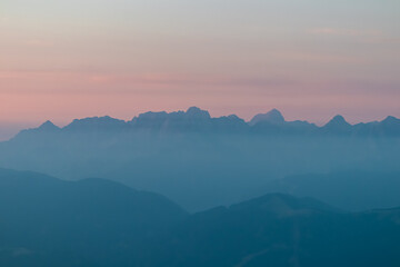 The majestic peaks of a distant alpine range emerge through layers of blue morning mist, viewed from Dobratsch. A serene and moody mountain landscape captured under a soft, pastel-colored dawn sky.