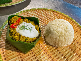 A traditional Cambodian Fish Amok, a yellow fish curry, is beautifully presented in handmade banana leaf bowl, topped with coconut cream and chili, served with portion of steamed rice on a woven tray