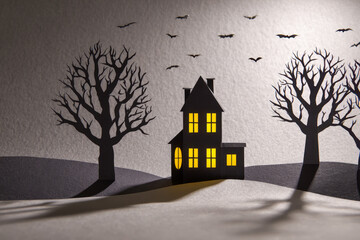 Paper cut out haunted house with glowing windows surrounded by barren trees and flying bats casting shadows on textured surface at dusk