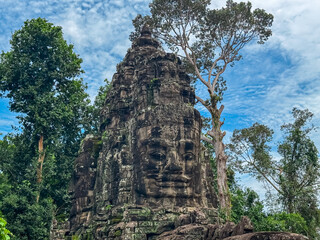 A detailed view reveals the intricate carvings and multiple serene stone faces of a tower at a gate to Angkor Thom, standing amidst lush green trees under a bright cloudy sky in Siem Reap, Cambodia.