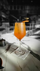 A refreshing orange cocktail, likely an Aperol Spritz, in a wine glass with a straw and orange peel garnish, resting on a white marble table in a stylish bar setting