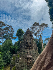 Ancient stone gate of Angkor Thom, adorned with serene faces, is framed by lush jungle canopy and a massive tree trunk under picturesque cloudy blue sky in Siem Reap, Cambodia's famous temple complex.