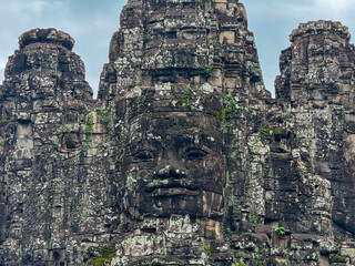 A detailed close-up shows the serene, smiling, and weathered stone face of Avalokiteshvara, a famous carving on a tower of the ancient Bayon temple in the Angkor Thom complex, Siem Reap, Cambodia.