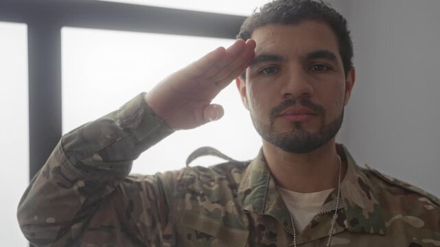 Man in military uniform salutes in a sunlit living room beside a large window while dog tags hang over his camouflage shirt and expression is resolute; duty.