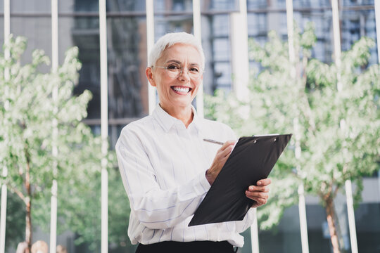 Confident elderly woman in a white striped shirt holds a clipboard outdoors in a modern city setting for business and professional use