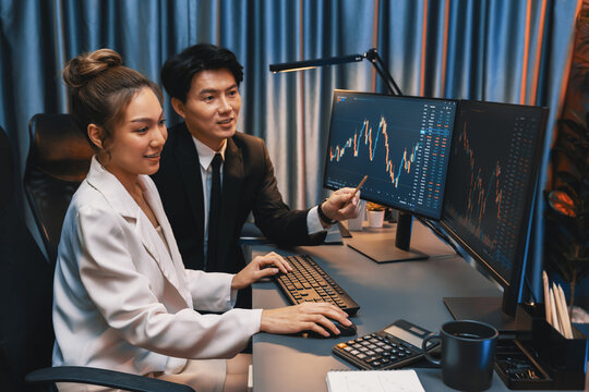 Smiling Asian business traders pointing to dynamic stock exchange data screen profit value online website program at night time at home office. Analyzing currency rate value on data report. Infobahn. - Powered by Adobe