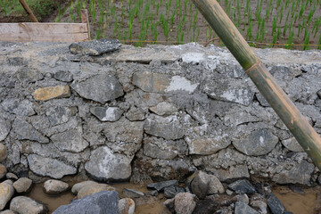 A close-up view of a house foundation made from stacked natural stones and cement, showing the rough texture and early construction stage on a muddy worksite.