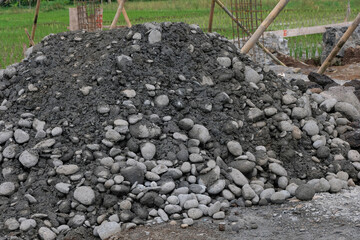 A pile of construction materials, including gravel and soil, at a house-building site with reinforced steel columns and wooden supports visible in the background.
