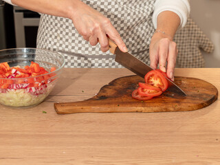 A woman slices a fresh tomato on a wooden cutting board while preparing ingredients for a homemade salad. Meal preparation step.