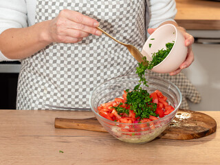A woman adds chopped parsley into a bowl with sliced tomatoes and vegetables. The process of preparing a fresh homemade salad.