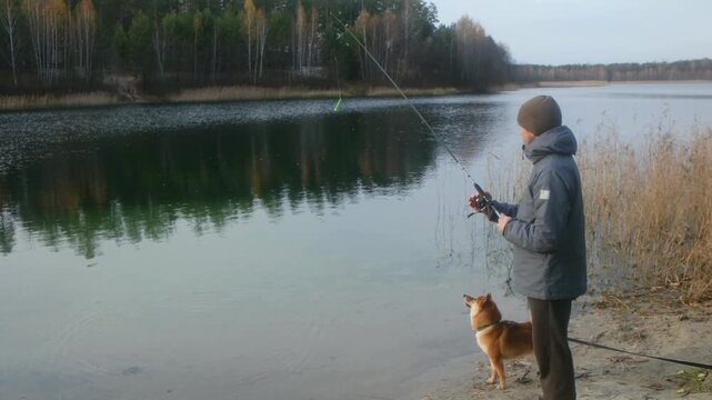 Fisherman standing on the sandy shore of a tranquil lake, casting a lure with a spinning rod while his faithful shiba inu dog watches attentively during a peaceful autumn day outdoors