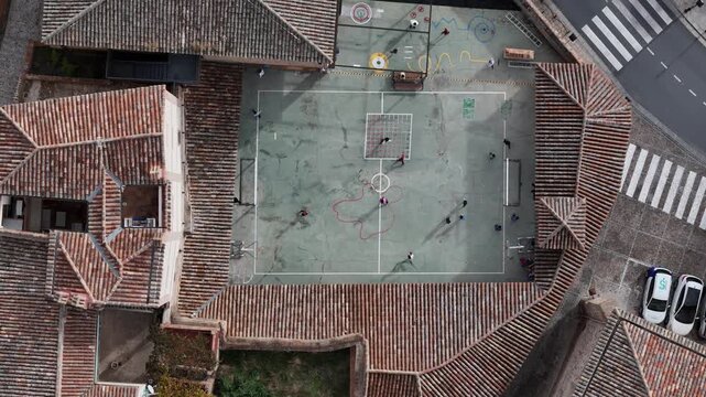 Top down aerial drone view of children playing on a colorful football court in a historic neighborhood during sunny afternoon hours