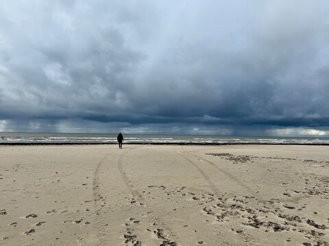 Solitary person walking along a wide sandy beach under dramatic storm clouds by the sea, creating a moody coastal atmosphere. - Powered by Adobe