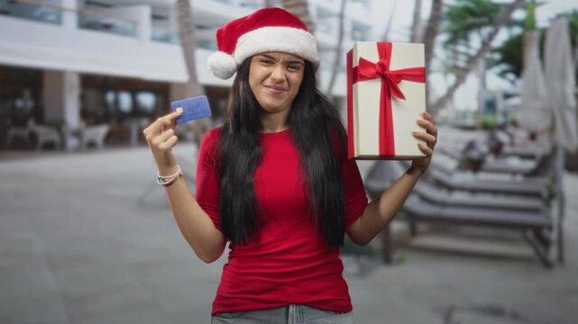 Young woman wearing santa hat and red shirt holds a creditcard and wrapped gift box at a resort poolside, holding card up and showing present; holiday shopping joy.