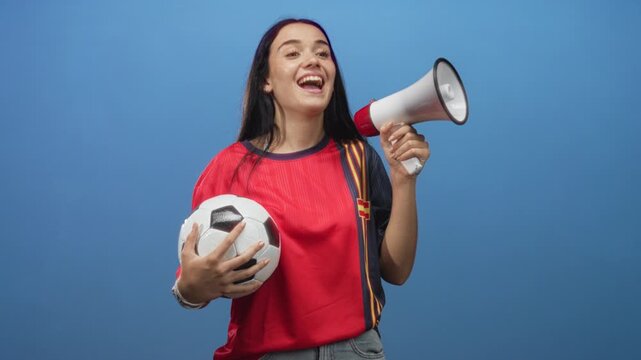 Woman wearing red team jersey holding soccer ball and megaphone, laughing and shouting in studio with blue backdrop; excitement.