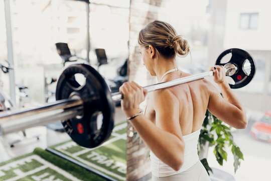 A fit woman is lifting a barbell in a modern gym, viewed from behind. Her back muscles are visibly engaged as she prepares to perform a squat or strength-training movement.
 - Powered by Adobe