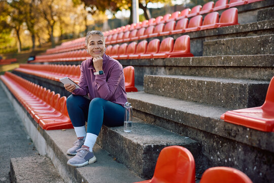 Mature athletic woman listening music over earbuds while relaxing on staircase during outdoor workout.