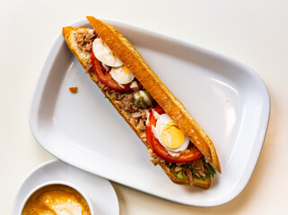 Spanish sandwich with tuna, egg and vegetables on fresh bread. Traditional baguette bocadillo con atun on a white plate side view