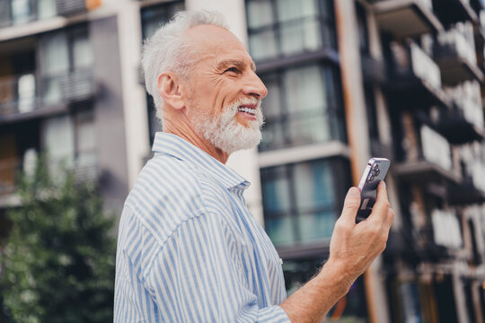 Senior man with white hair and beard using smartphone in urban cityscape smiling outdoors among modern residential buildings during daytime