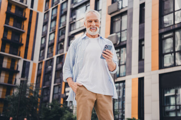Senior man in casual striped shirt smiles using phone outdoors amid modern city architecture urban...