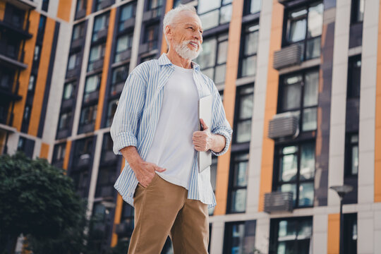 Senior man stands outdoors beside a modern residential building holding a tablet and smiling in a contemporary urban city scene - Powered by Adobe