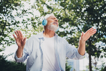 Joyful elderly man with blue headphones enjoying outdoor music in a sunny city park during a lively...