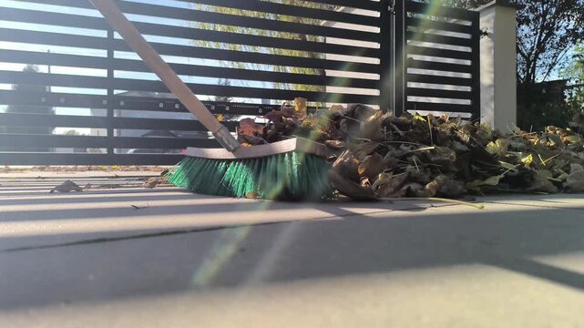 Zamiatanie opadłych suchych jesiennych liści z podjazdu szczotką o zielonym włosiu.  Czyszczenie podjazdu szczotką z syntetycznego włosia. W tle widoczna brama wykonana z poprzecznych belek stalowych.