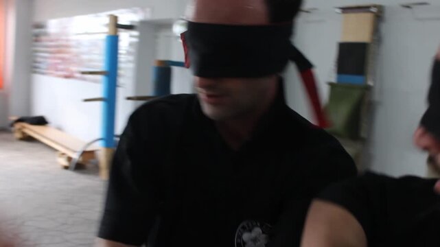 Two martial artists wearing black uniforms and red blindfolds practice Wing Chun Chi Sao sticky hands drills to improve their tactile sensitivity and reflexes inside a training studio.

