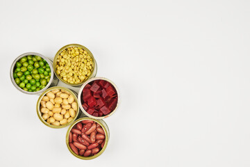 Open cans of beans, mushrooms, carrots, corn, peas, green beans, lentils, asparagus, and olives on a white background with copy space.