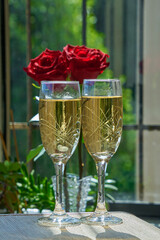 Two champagne glasses with red roses against a backdrop of green foliage for a romantic celebration or Valentine's Day.