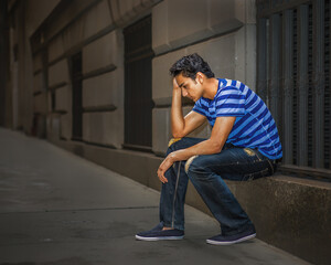 Pensive Young Man in Urban Setting. A young man sits on a stone ledge with a thoughtful expression. The street is deserted, adding to the contemplative atmosphere.