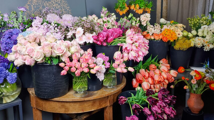 Colorful assortment of fresh flowers displayed in black buckets inside flower shop. Various blooms including tulips, roses, mixed spring flowers arranged on and around a wooden table on floral market 