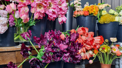 Colorful assortment of fresh flowers displayed in black buckets inside a flower shop. The arrangement includes pink tulips, purple parrot tulips, orange blooms, and various seasonal flowers 