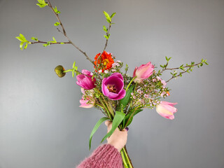 Hand holding a spring bouquet with pink tulips, blooming branches, and delicate wildflowers against a neutral gray background. Florist work at flower shop