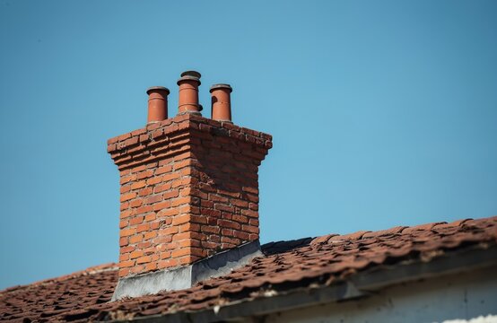 Old red brick chimney with three clay pots on a weathered tile house roof. Traditional building rooftop against a clear blue sky. Architectural detail of a vintage home exterior.