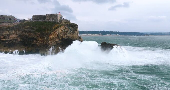 Aerial drone shot of Farol da Nazare lighthouse during a big wave professional surfing event in Nazare, Portugal, Europe. Drone orbit, waves crashing into cliff, spectators watching