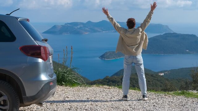 Tourist raising arms enjoying scenic view from mountain road. Joyful traveler exiting vehicle and stretching arms, celebrating scenic mountain road overlooking vast ocean with excitement and freedom