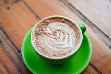 An eye-level shot features a cappuccino in a bright green cup with a matching saucer. The creamy coffee has a leafy design. The image evokes warmth and relaxation on wood background.