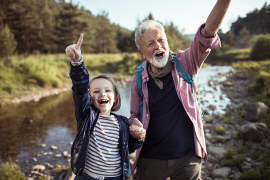 Senior grandfather and child granddaughter cheering while hiking by forest creek