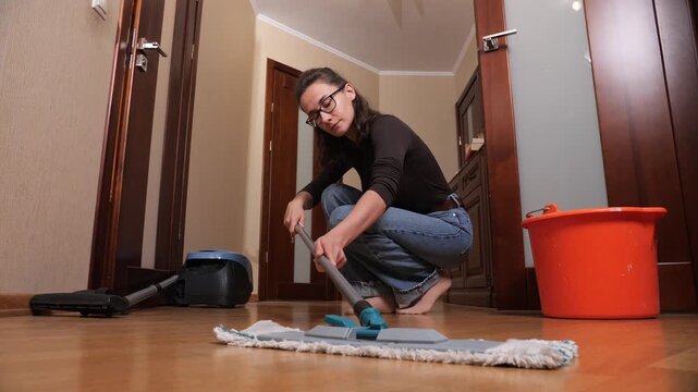 Woman mopping a light wooden floor at home, wearing casual clothes and glasses, diligently cleaning the floor with a flat mop and bucket in a residential hallway