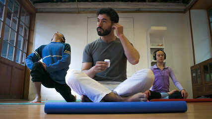 A bearded man sits cross‑legged on a blue mat and takes earphones, while two others stretch and breathe at back.