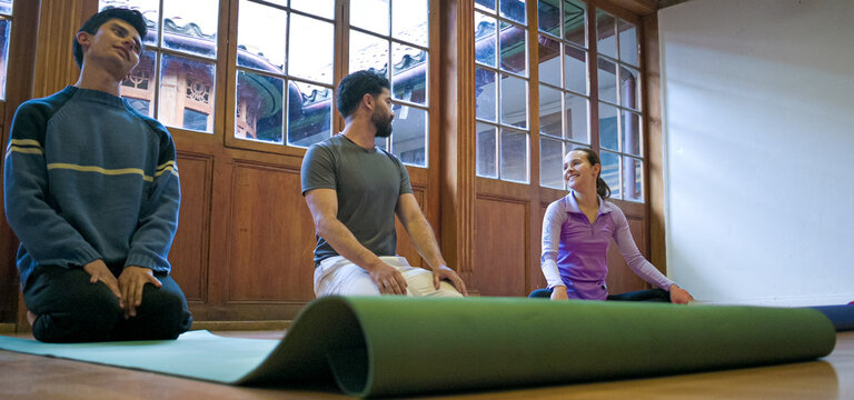 Low angle view of three friends sit on colored yoga mats in a bright studio with wooden windows, sharing a light moment during a group yoga session.
