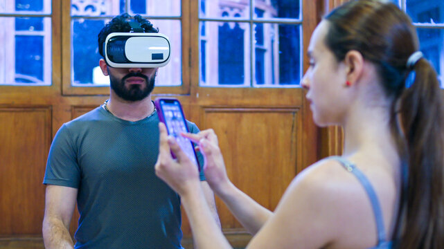 Woman nearby interacts with a smartphone, while man wears a virtual reality headset immersed in a digital relaxing experience at background.
