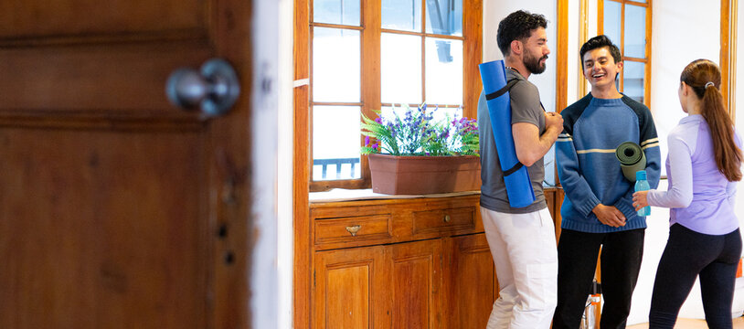 A group of friends talking and smiling during a casual indoor fitness break, holding yoga mats and a water bottle near a wooden cabinet.  - Powered by Adobe