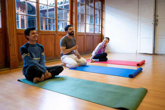 Three friends sit on colored yoga mats in a bright studio with wooden windows, sharing a light moment during a group yoga session. - Powered by Adobe