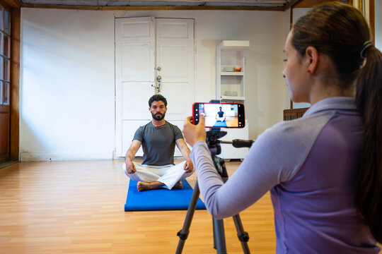 A focused man sits cross-legged on a blue yoga mat in a bright studio as a woman records him with a smartphone on a tripod, capturing a calm mindfulness moment.