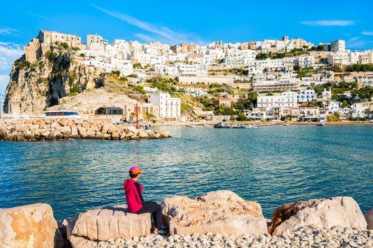 Female traveler sitting on coastal rocks admiring the whitewashed town of Peschici overlooking the sea, Apulia, Italy.