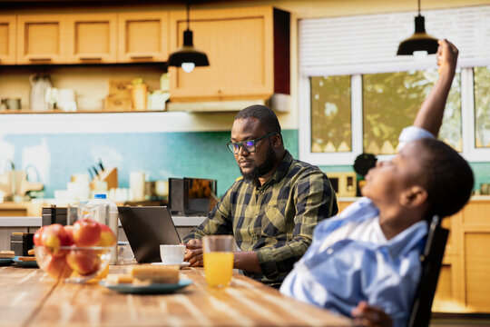 Father working on his laptop and serving breakfast with his cute family, gathered in the kitchen to enjoy the simple comfort of morning togetherness. Happy start to the day with sunlight.