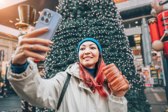 Happy young woman taking selfie with a smartphone and authentic Trdelnik at snowy European Christmas market