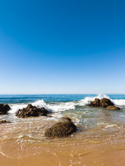 Waves crashing on rocks on a sandy Southern California beach under a vibrant clear blue sky